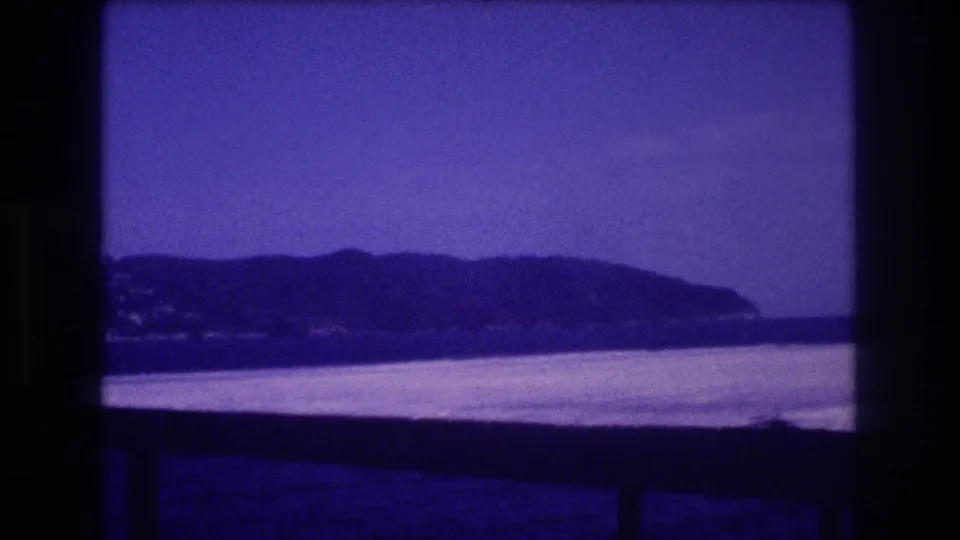 Home-movie frame from the supplied Acapulco clip showing a low, hazy shoreline beyond water, partly obscured by the boat edge and film frame.