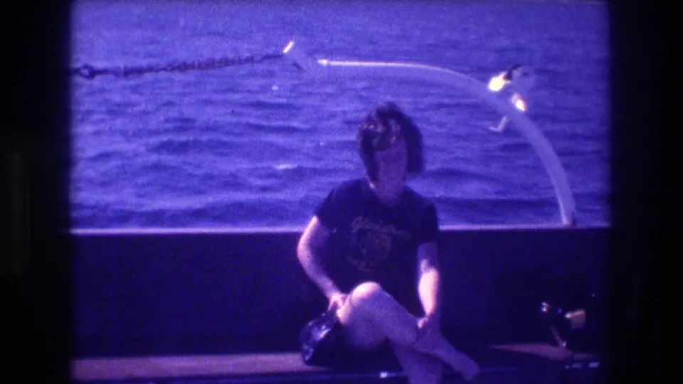 Blue-tinted 8mm frame from the supplied 1982 Acapulco footage showing a dark-haired woman seated on a boat with open water and a curved boat fixture behind her.