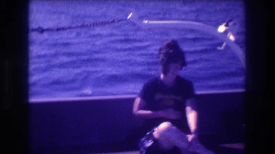 A dark-haired woman sits low on the boat, appearing uncomfortable as she holds her midsection while the blue sea and rail remain behind her.