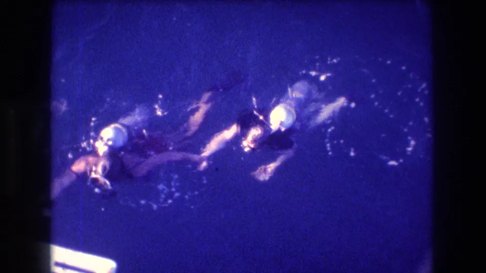 Two scuba divers in masks and fins swim underwater in dark blue sea, leaving bubbles as they move through the frame.