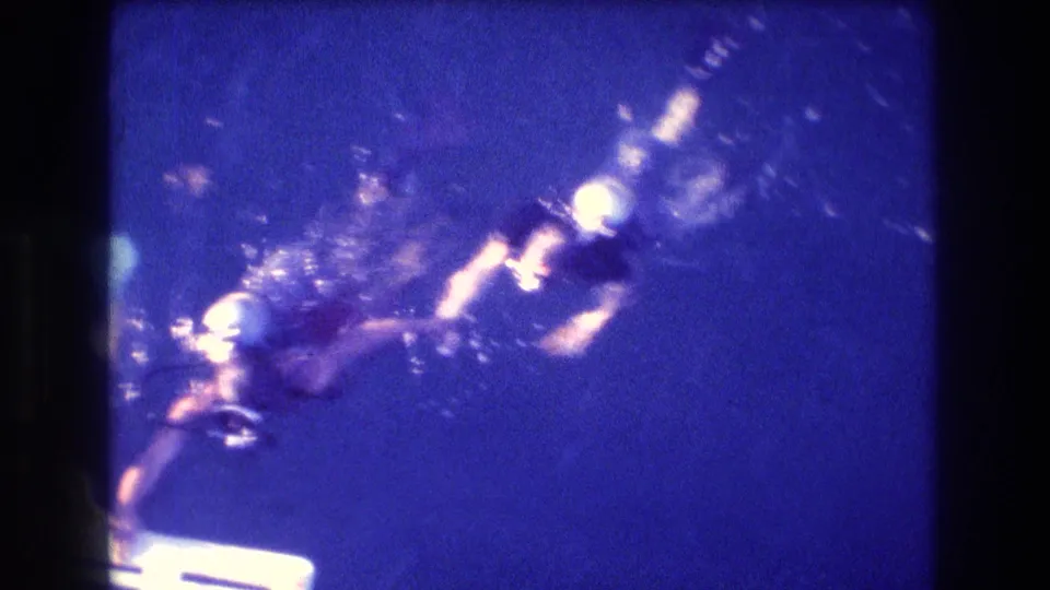 A scuba diver in mask, tank, and fins moves through blue water near a white ladder or platform in 1982 Acapulco home-movie footage.