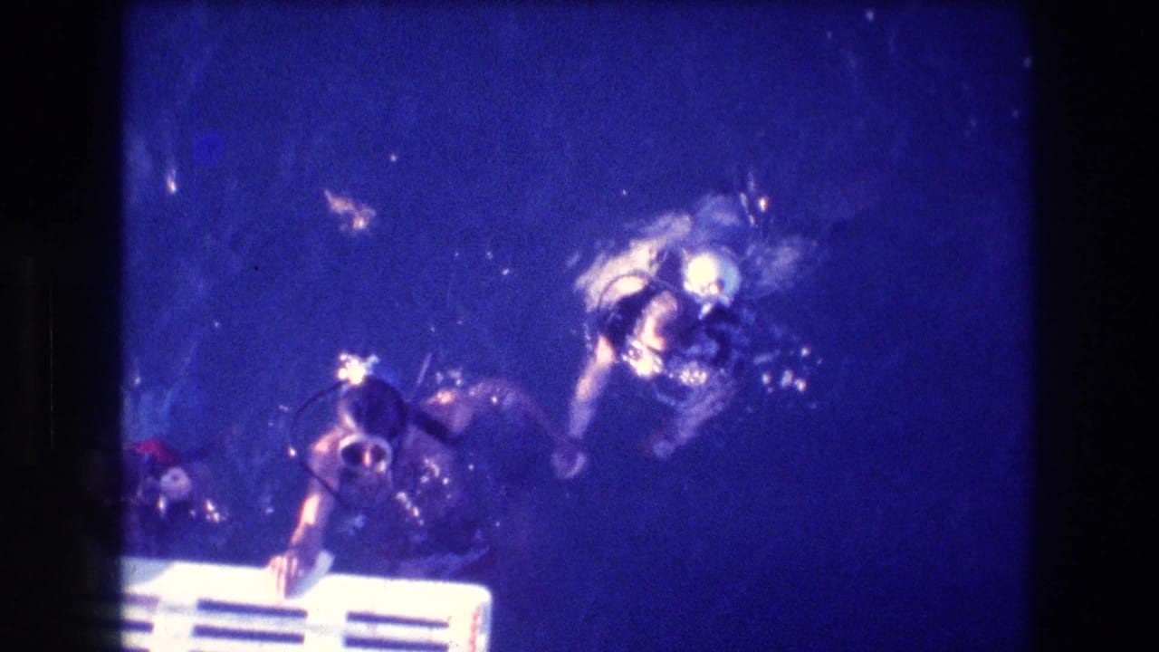 Two scuba divers in masks and tanks approach the surface area above a white ladder or platform in blue underwater 8mm footage from Acapulco, Mexico.