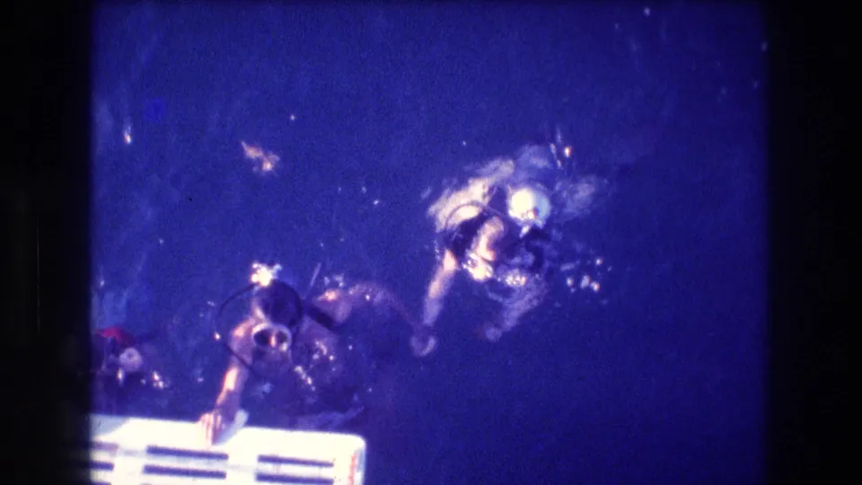 Two scuba divers in masks and tanks approach the surface area above a white ladder or platform in blue underwater 8mm footage from Acapulco, Mexico.