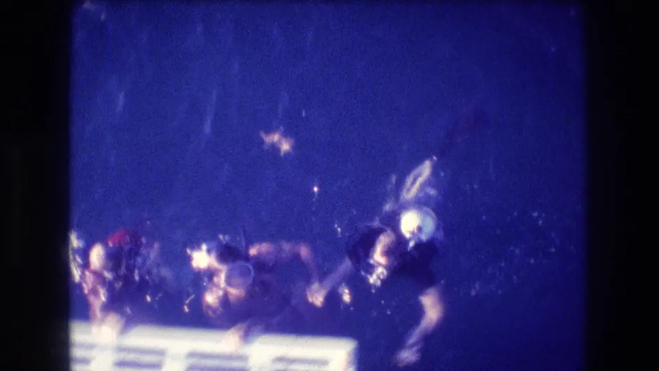 Several scuba divers in masks and tanks cluster near a white platform or ladder in grainy blue 8mm home-movie footage from Acapulco in 1982.