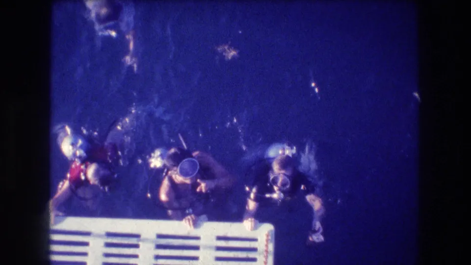 Multiple scuba divers in recreational gear surface around a white ladder or platform, with bubbles and heavy blue underwater color in 1982 Acapulco 8mm footage.