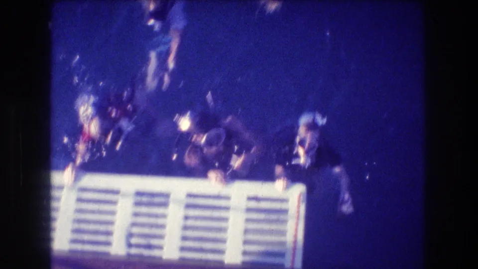 In 1982 Acapulco home-movie footage, divers wearing masks and snorkels cluster near a white slatted structure in deep blue ocean water.