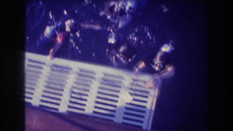 A group of divers in dark gear holds onto a white slatted structure in ocean water, captured with soft 8mm grain and strong blue color shift in Acapulco, Mexico.
