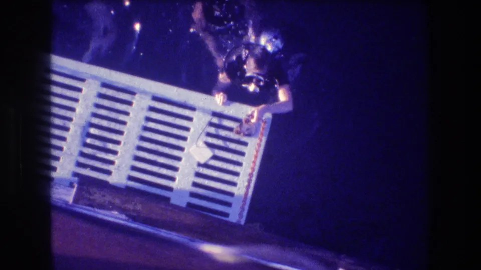 A diver wearing a mask and dark gear holds the end of a white slatted structure in blue ocean water, with other swimmers partly visible nearby in the 1982 Acapulco footage.