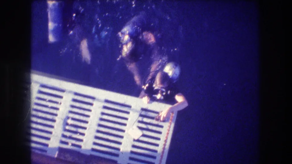 In 1982 Acapulco home-movie footage, two scuba divers hover beside a white slatted underwater platform in deep blue water.