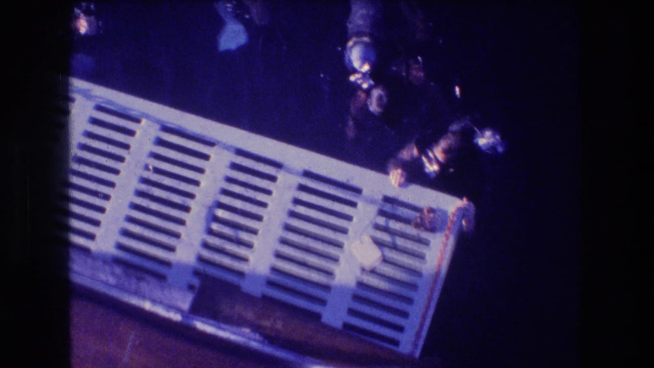 Two scuba divers in masks and tanks hold close to a white slatted underwater platform, seen through blue 8mm film color shift.
