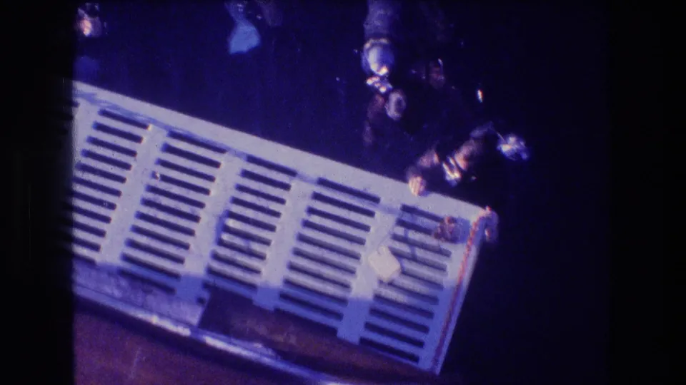 Two scuba divers in masks and tanks hold close to a white slatted underwater platform, seen through blue 8mm film color shift.