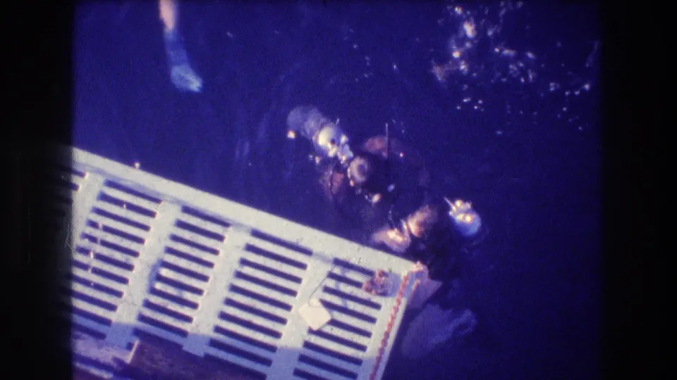 The underwater home-movie frame shows divers near the corner of a white slatted platform, with bubbles and heavy blue exposure.