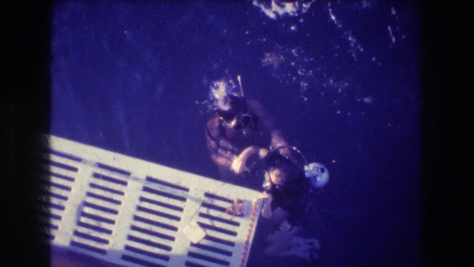 Two scuba divers remain beside the white slatted platform in deep blue water, framed as vintage 8mm Acapulco travel footage from 1982.