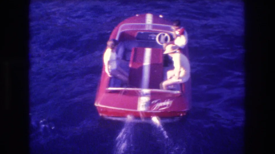 High-angle 1982 Acapulco home-movie frame of a red speedboat with white stripes carrying three people over dark blue ocean water.