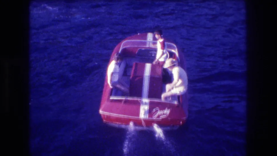 1982 Acapulco home-movie frame of three people aboard a red speedboat, including a seated driver and rear passenger, as the boat leaves a bright wake.