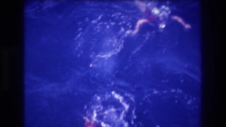 A swimmer is partly visible in the blue ocean, surrounded by bubbles, wake patterns, and soft 8mm film blur from the elevated home-movie perspective.
