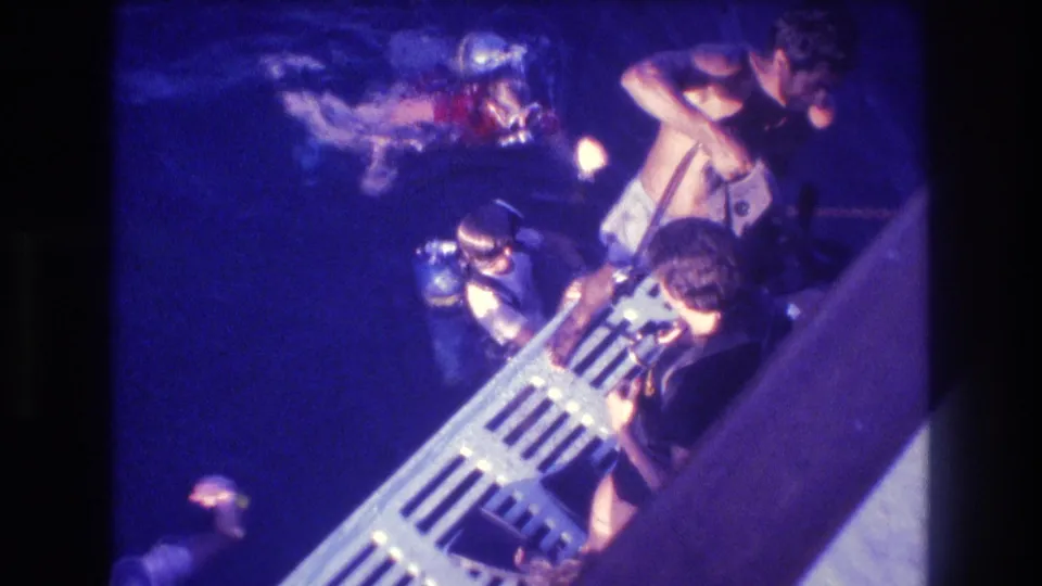 Overhead 8mm shot from a dive boat circa 1982, showing a small group near a metal boat railing and ladder over deep blue ocean water.