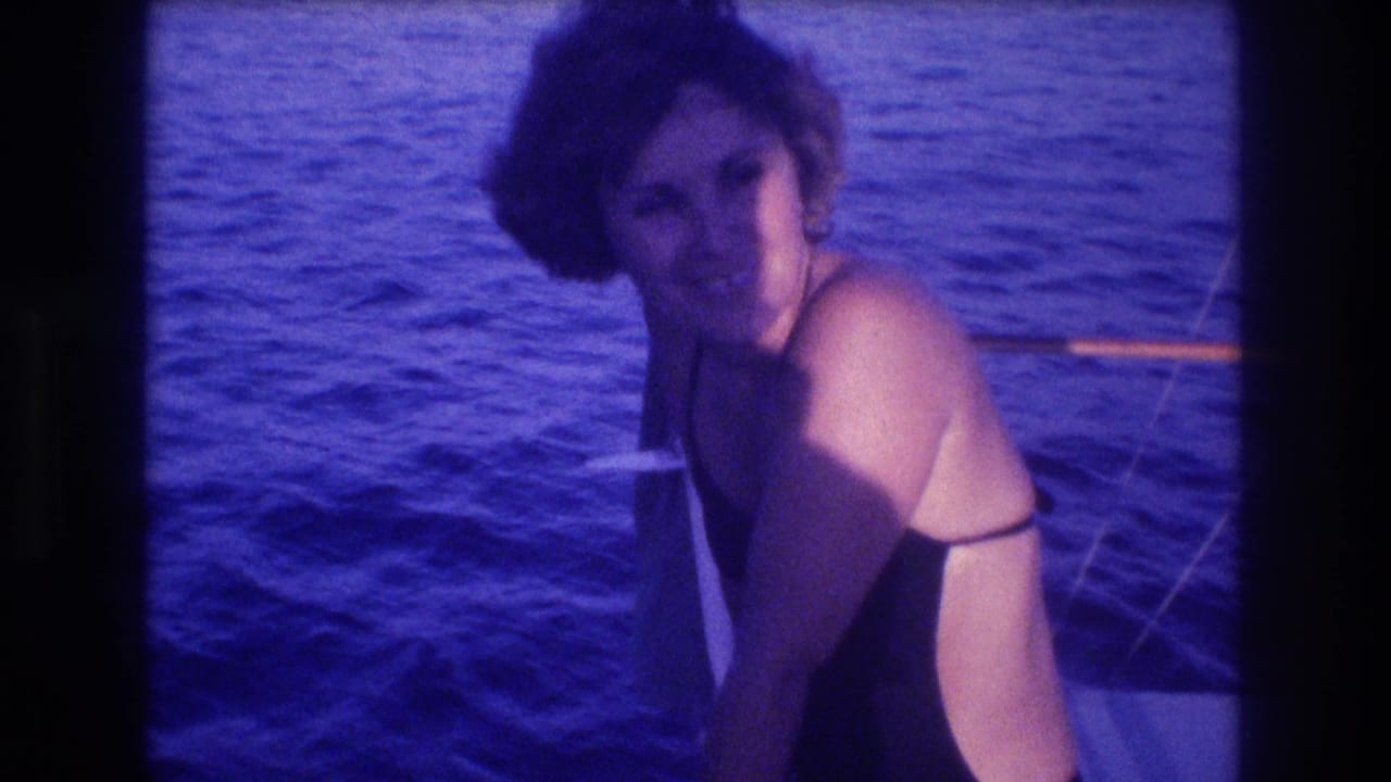 Woman in dark one-piece swimsuit smiling at camera on boat deck, choppy blue ocean behind, strong blue-purple 8mm color cast.