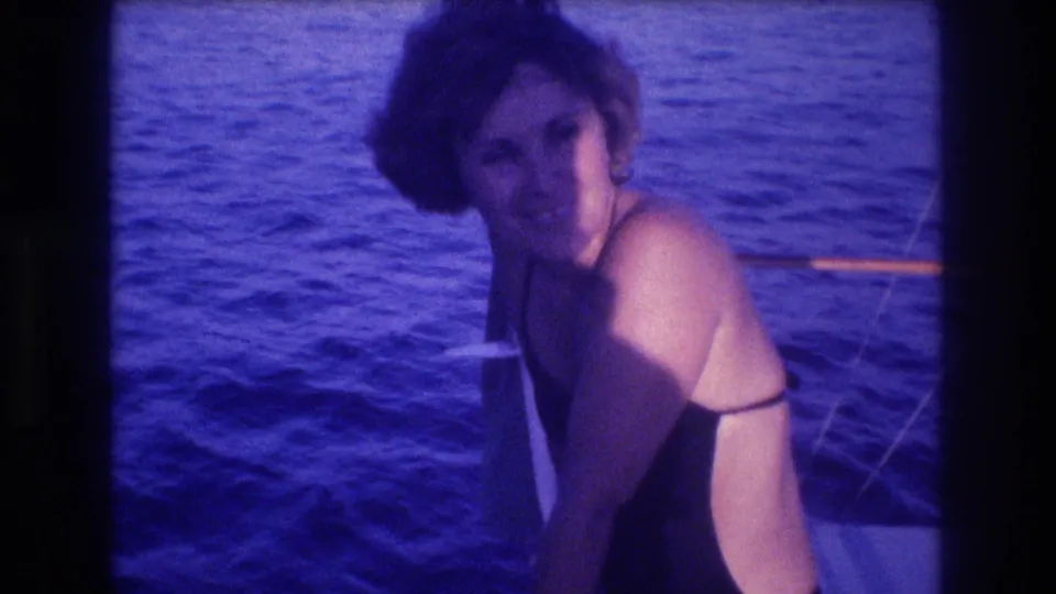 Woman with short wavy hair in dark one-piece swimsuit smiling at camera on a boat, blue ocean behind, 8mm home movie, Acapulco, Mexico, circa 1982.