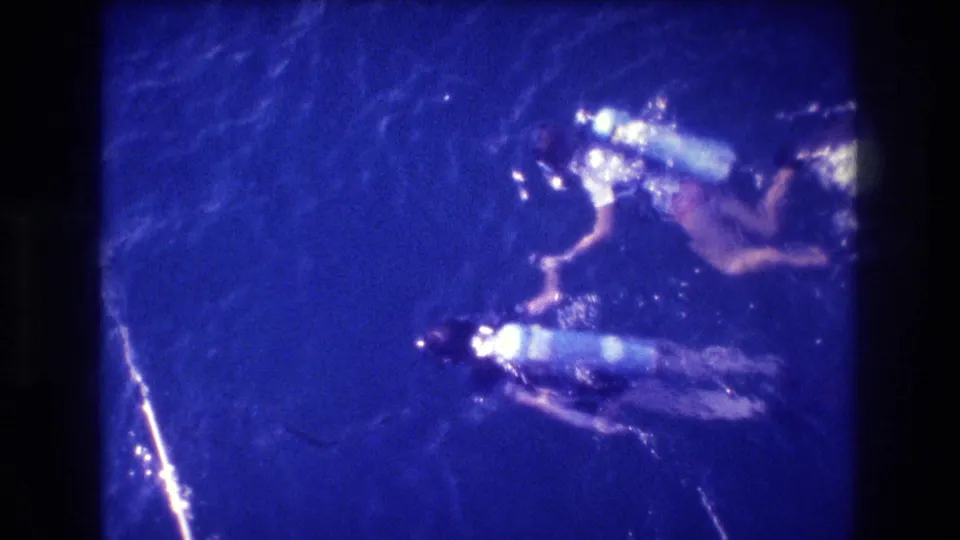Overhead view of two scuba-equipped figures in blue-shifted 8mm footage, with a strong water-surface reflection; consistent with 1982 home-movie material per clip metadata.