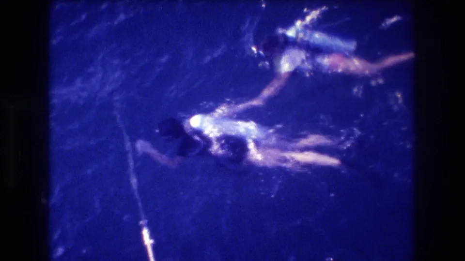 Overhead shot of two swimmers, one in lighter attire with scuba tank, and a vertical rope or line at lower-left; blue-shifted 8mm stock consistent with early-1980s home-movie material.