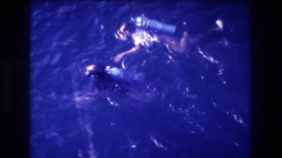 Close overhead view of two divers with visible air tanks in blue-shifted 8mm footage; composition consistent with prior frames from what metadata identifies as a 1982 Acapulco home movie.
