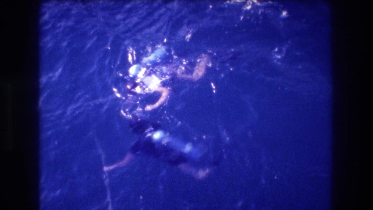 Overhead 8mm frame of two swimmers in blue-shifted ocean; a blue flotation device is visible near one figure, Acapulco area, 1982.