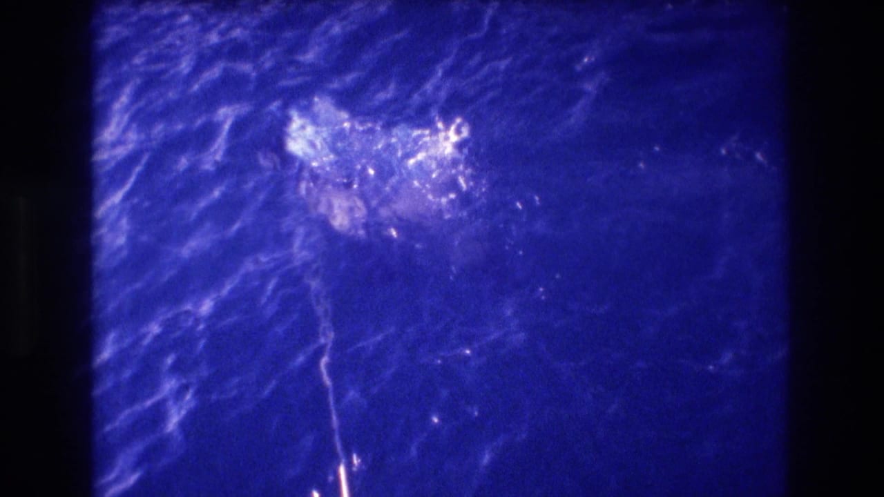 Continued shot of two people swimming in 8mm home movie footage with pronounced blue color shift; figures appear as partially submerged shapes in motion.
