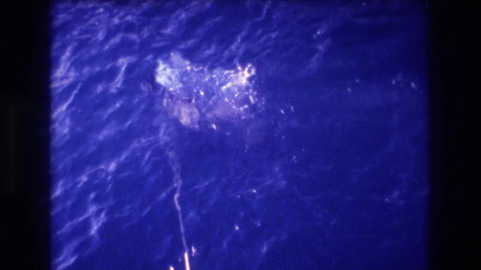 Continued shot of two people swimming in 8mm home movie footage with pronounced blue color shift; figures appear as partially submerged shapes in motion.