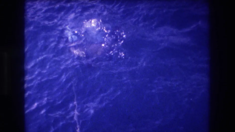 One swimmer visible prominently in 8mm home movie footage with heavy blue cast, reportedly Acapulco 1982; second figure barely discernible amid rippling water.