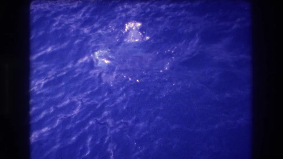Final frame of 8mm ocean home movie with heavy blue shift and grain dominant; a figure is faintly visible amid the rippling water surface.