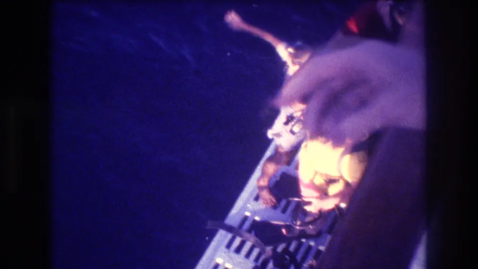 Super 8 home-movie frame showing a figure in yellow garment leaning over a boat's metal railing toward open ocean, likely Acapulco 1982.