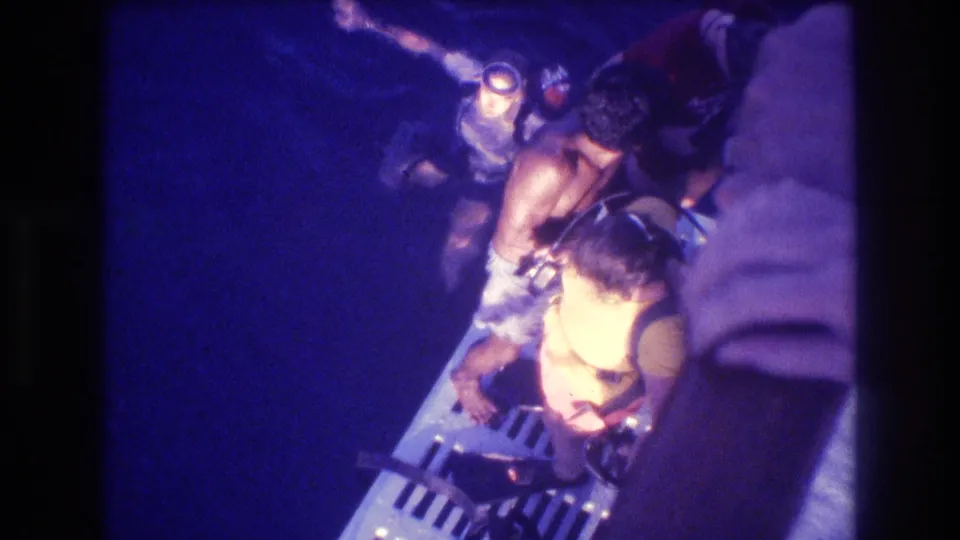 Closer overhead view of a child in a yellow life vest and adult on a boat platform at night, Acapulco, Mexico, circa 1982, 8mm home movie.
