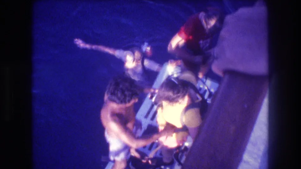 Overhead shot on a small dive boat, Acapulco Mexico 1982: a shirtless man and a person in a yellow top manage scuba equipment under artificial light against dark water.