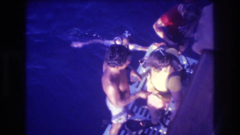 Closer overhead angle on a dive boat deck, Acapulco Mexico ca. 1982: shirtless man with dark hair assists another diver with scuba tank and regulator under warm artificial light.