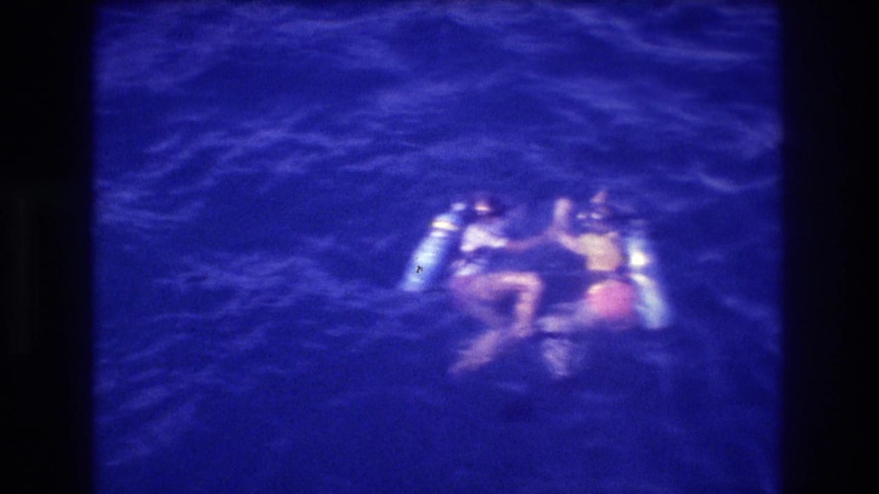 Two scuba divers floating on the ocean surface near Acapulco, Mexico, circa 1982, one with arm raised, on 8mm home movie film.