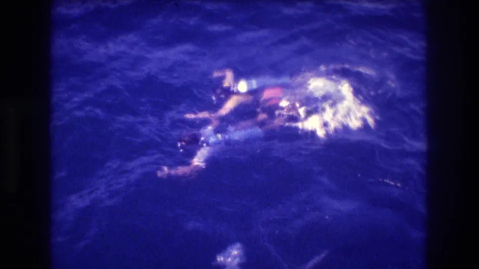 Lone swimmer at ocean surface with blue snorkeling gear and red-orange swimwear, overhead angle consistent with 1982 Acapulco vacation home-movie footage.
