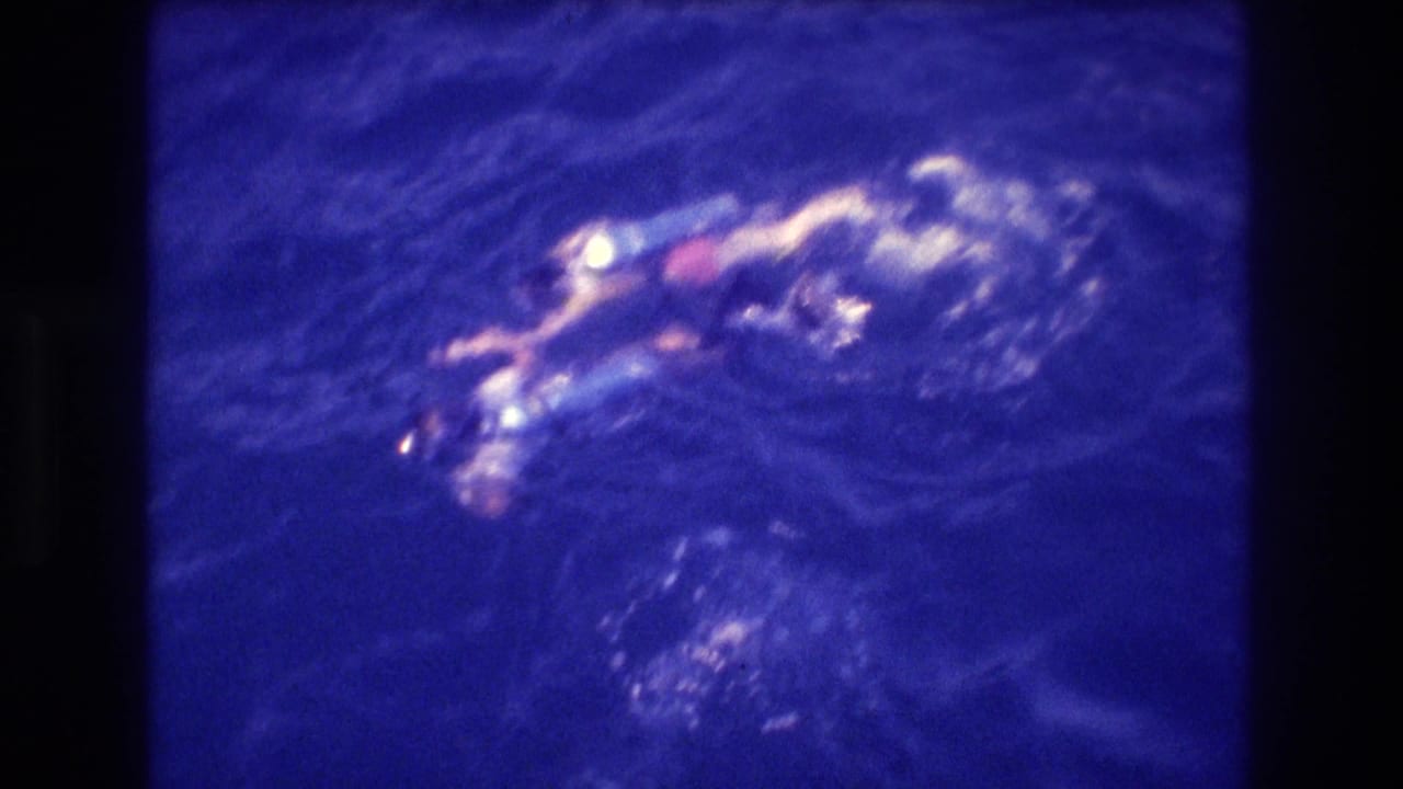 Overhead view of a swimmer at the ocean surface amid churning white water, shot from a boat with heavy blue color cast.