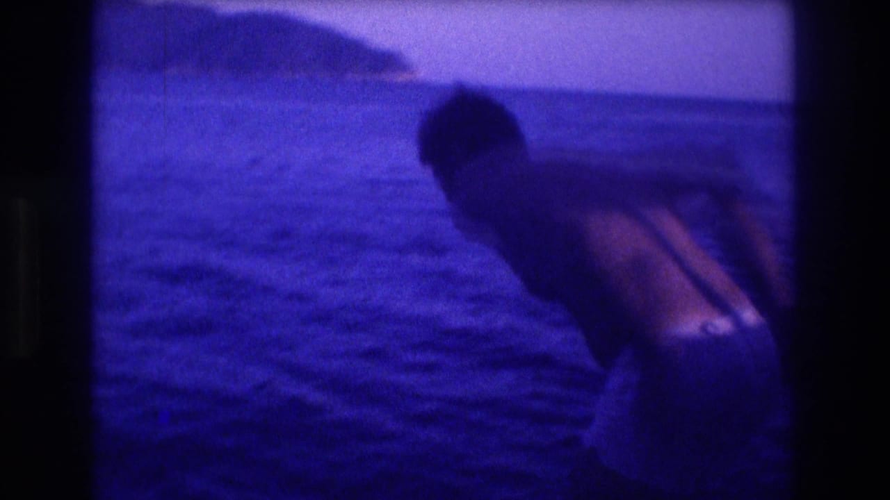 Person leaning over the side of a boat toward the water, with a rocky coastal headland visible in the background and strong blue-purple color cast.