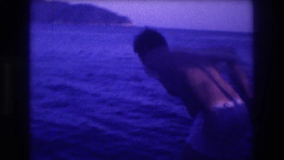8mm home-movie frame showing a figure leaning from a boat toward the sea, with a rocky headland behind; Acapulco area, 1982.
