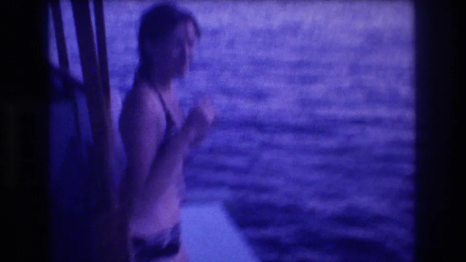 Adult in dark swimsuit at boat railing, partial profile view, with open ocean behind; 8mm home movie, circa 1982 Acapulco, Mexico; blue film cast and grain visible.