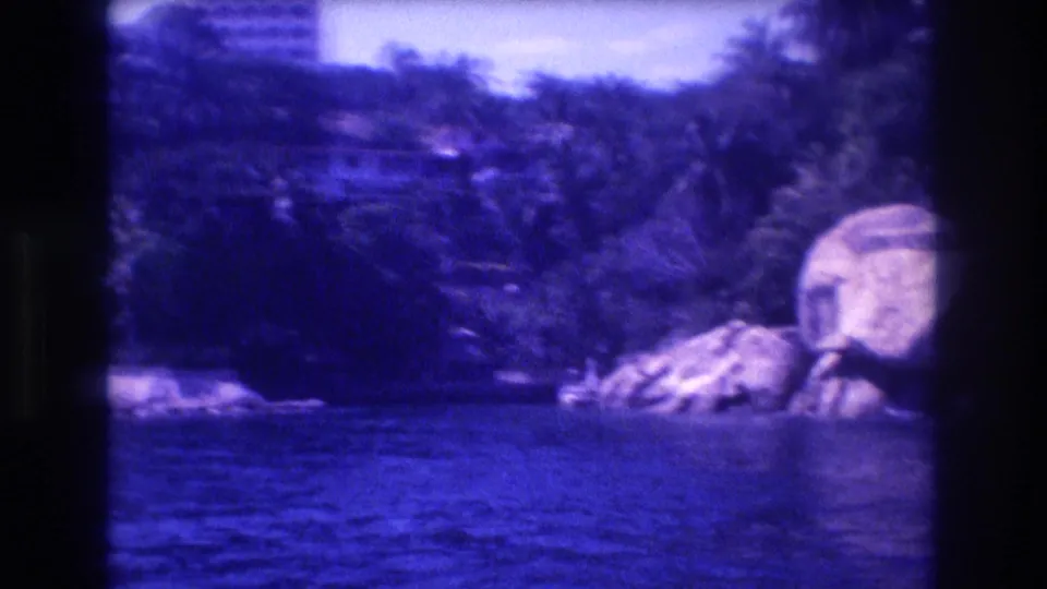 8mm view from a tourist boat of large boulders on the Acapulco shoreline with dense palms and a resort building visible above, circa 1982, with blue color drift.