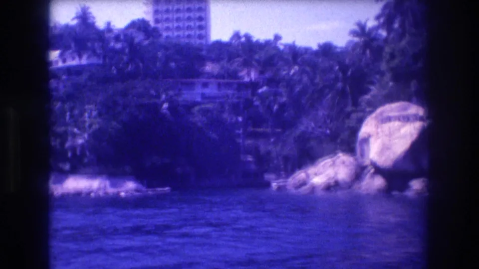8mm boat-ride view of the Acapulco shoreline showing rounded boulders, palm vegetation, and a high-rise resort building circa 1982, with heavy blue film cast.