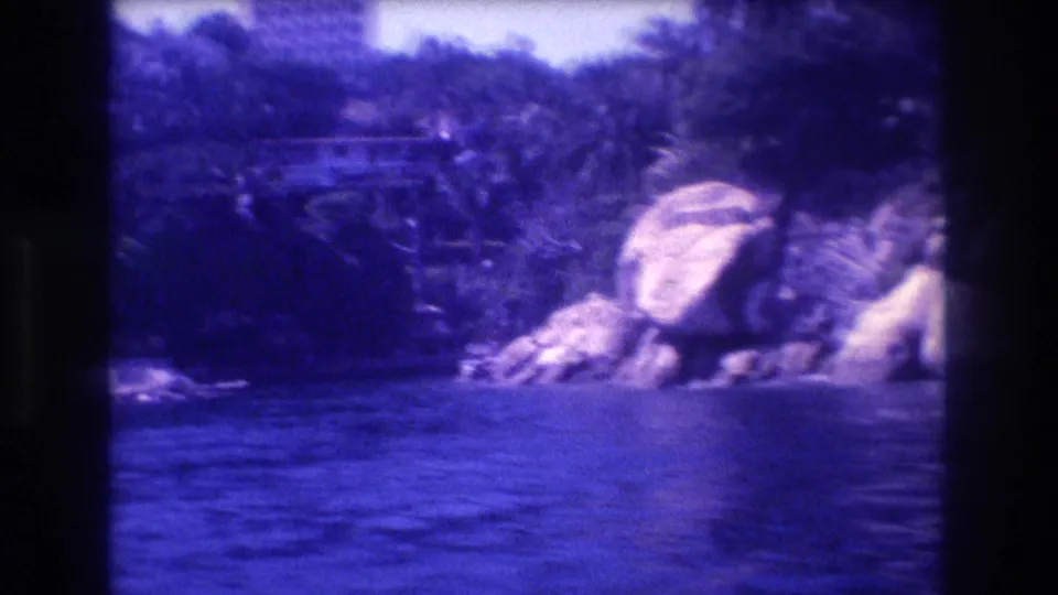 Rocky coastline likely in Acapulco, Mexico, circa 1982; large boulders, tropical palms, and a high-rise building in the background; 8mm blue-violet color cast throughout.