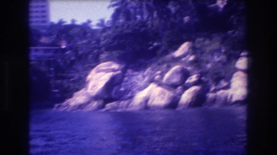 Acapulco coastline, likely circa 1982, from a moving boat; rounded boulders at the waterline, lush tropical palms, and a prominent high-rise building at upper right; 8mm film.