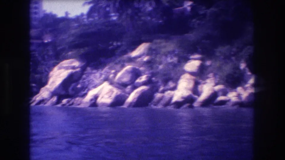 Boulder-strewn coastline, likely Acapulco, Mexico, circa 1982; large rocks crowd the waterline with calm bay water below and tropical vegetation above; 8mm color cast.