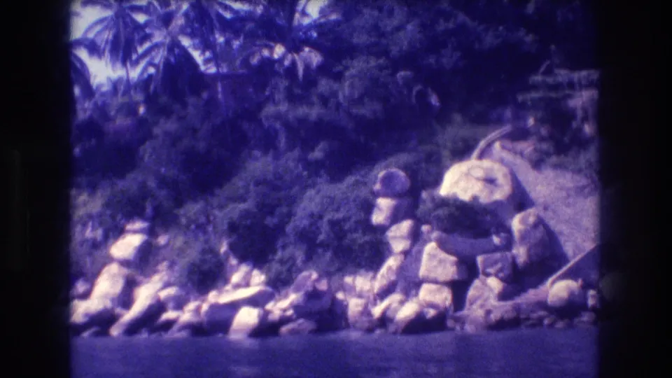 Acapulco, Mexico, 1982 — boulders along a tropical waterway with palm trees, captured on 8mm home movie film with characteristic purple aging shift.