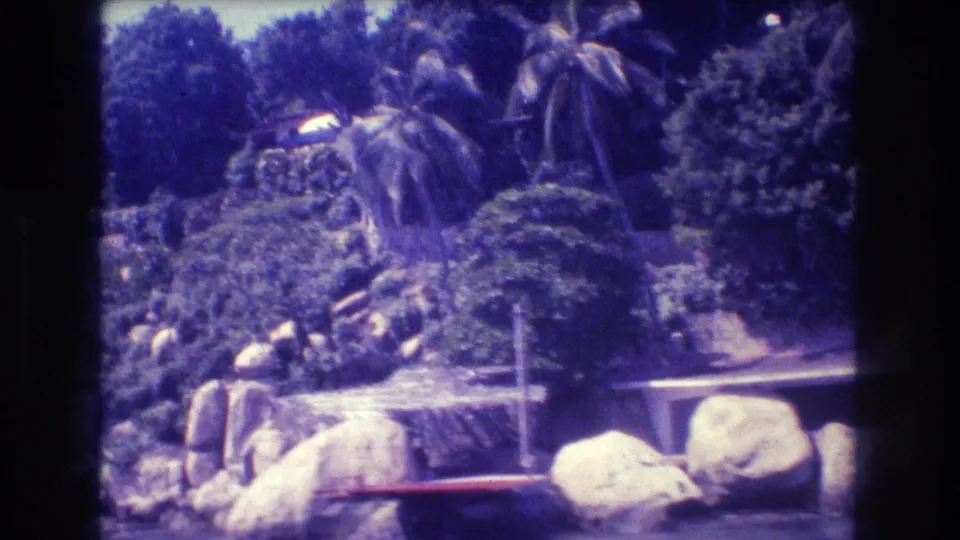 Acapulco, Mexico, 1982 — final pan position showing boulder-strewn tropical waterway with palm trees and partially visible background structures on aged 8mm film.