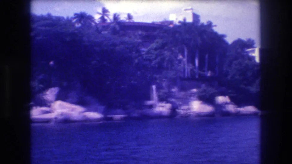 8mm boat-level view of a rocky tropical headland covered in palm trees, with a faint white structure glimpsed through the canopy, Acapulco area circa 1982.