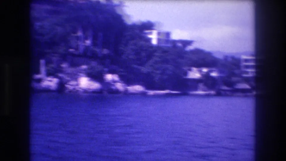 Slightly blurred 8mm frame showing a multi-story building above the rocky hillside trees, shot from a moving boat off Acapulco, circa 1982.